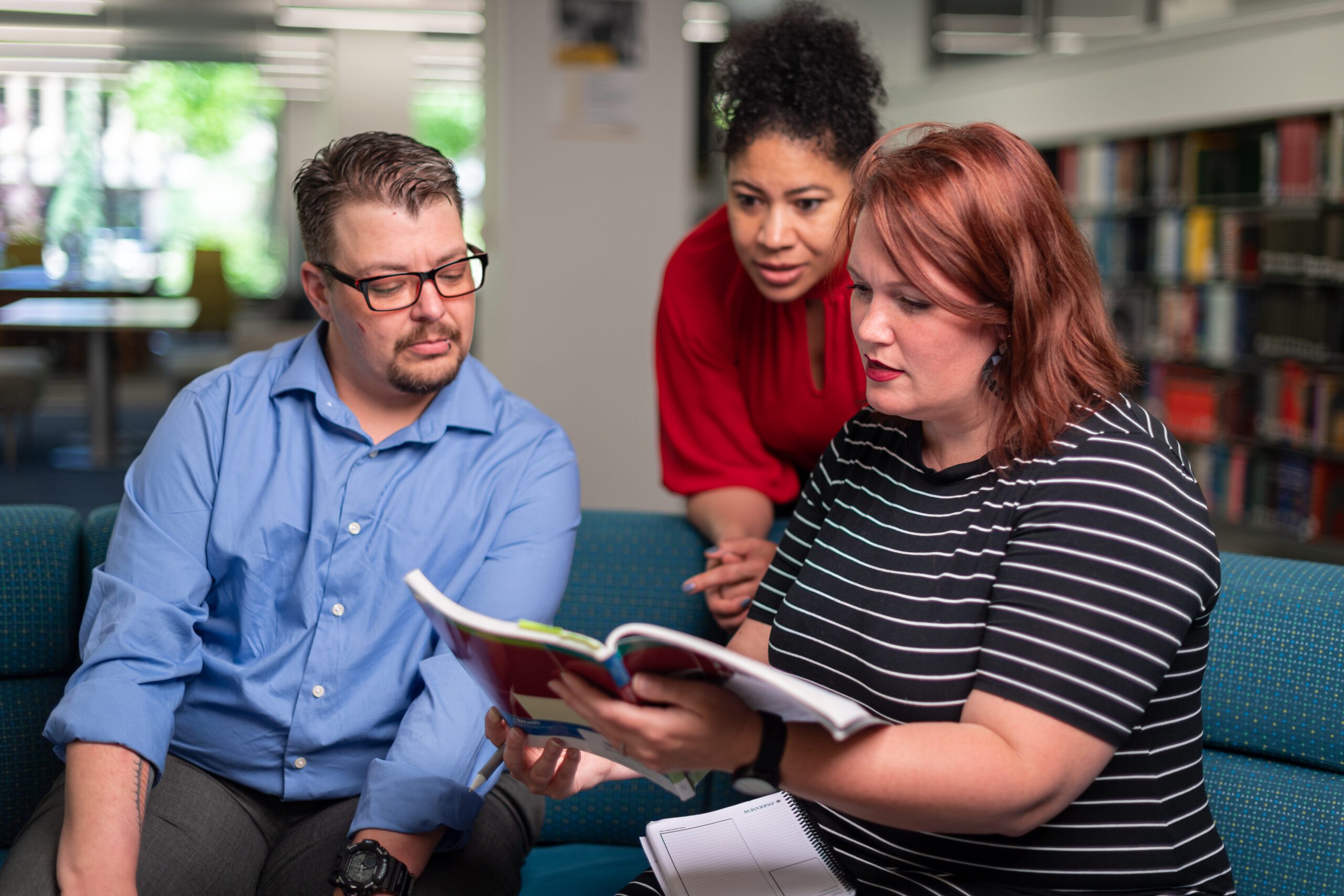 Three people sit in a seating area in a library and overlook a workbook together